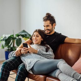 Ein Paar liegt auf dem Sofa, gemeinsam schauen sie auf ihr Smartphone.